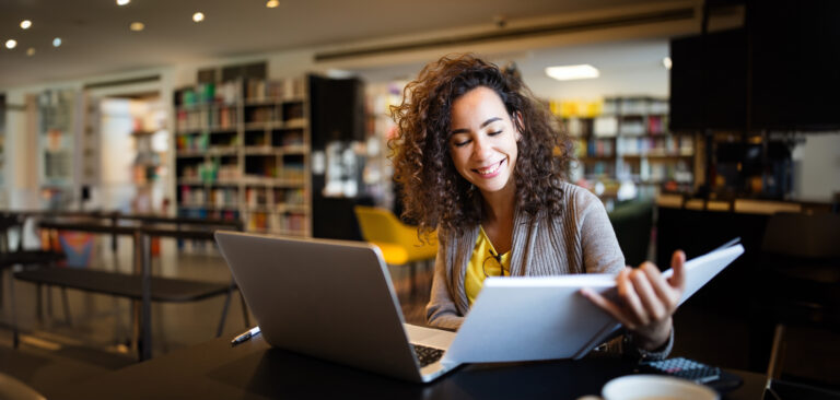 smiling young woman working at a desk with her laptop and a book